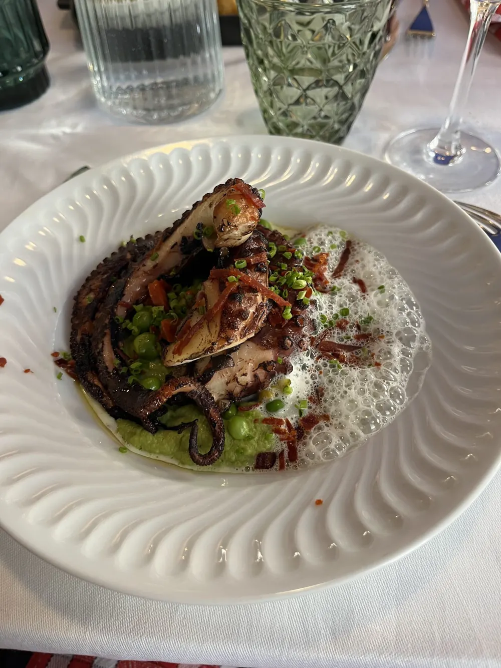 Poulpe grillé sur une purée verte avec des petits pois et une mousse blanche, dans une assiette blanche cannelée